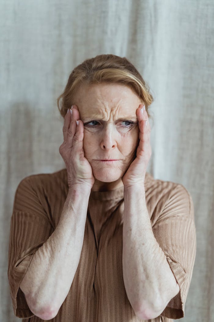 services-05 Portrait of an elderly woman showing stress and concern with hands on face indoors.